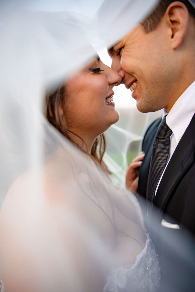 A bride and groom smile under a veil. 